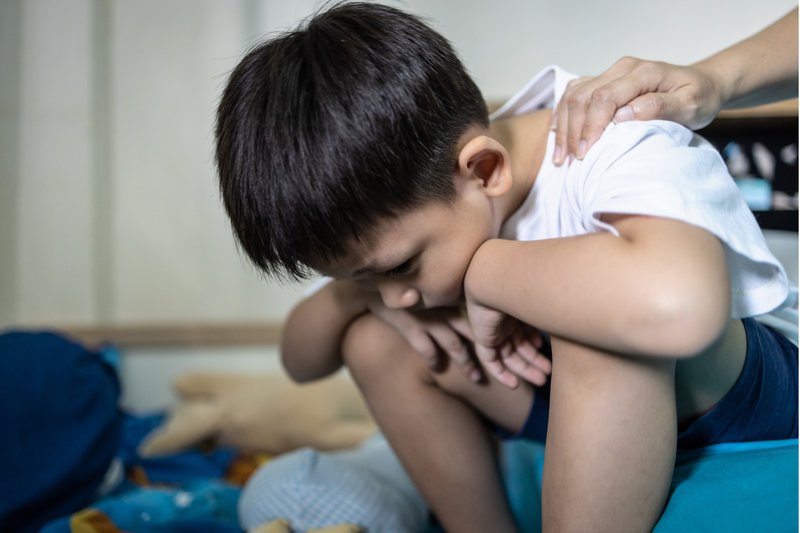 Jonge kind met zwart haar en witte t-shirt. Hij zit voorovergebovgen en lijkt verdrietig. Op zijn schouder zien we een bemoedigdende hand van een vrouw. De vrouw is niet in beeld, we zien enkel haar hand en arm.