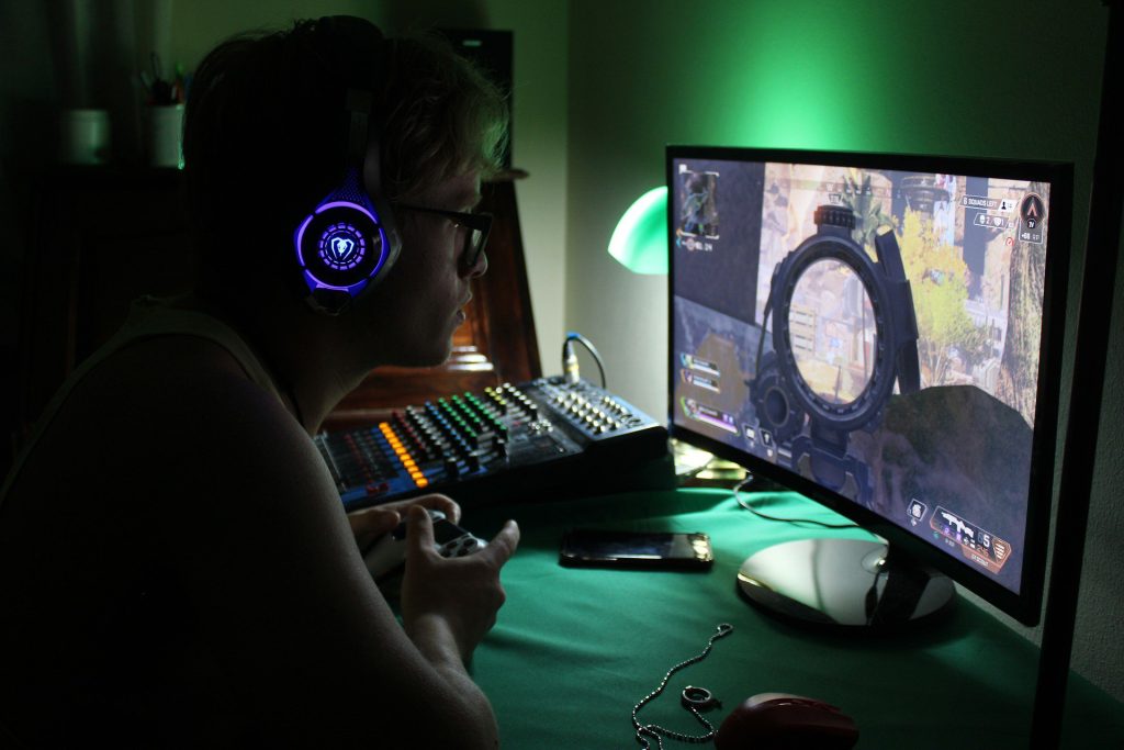 Jongvolwassen kind die naar een computerscherm kijkt. Het kind zit in het donker, heeft een bril op en een koptelefoon die licht geeft. Hij heeft een gamecontroller in de hand, en speelt een spel op de computer.Op het scherm zien we een visual die doet vermoeden dat het kind een actiespel speelt.