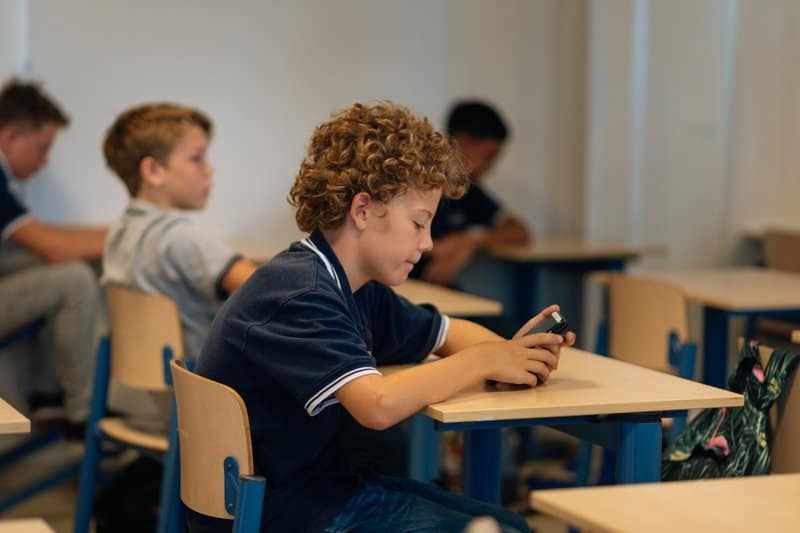 Een jonge man zit ontspannen op een bureaustoel, met zijn voeten op het bureau en kijkt naar zijn telefoon.