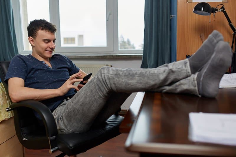 Een jonge man zit ontspannen op een bureaustoel, met zijn voeten op het bureau en kijkt naar zijn telefoon. Door het smartphoneverbod is er mogelijk meer schermtijd thuis.