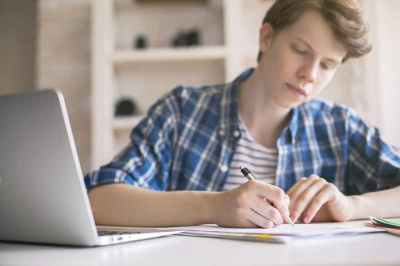 Een jonge student met blond haar en een geruit overhemd schrijft geconcentreerd in een notitieboekje, met een open laptop links op tafel, wat de balans tussen traditioneel leren en technologie symboliseert.