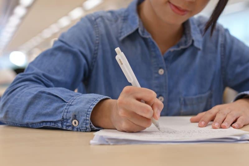 Close-up van een persoon die met een witte pen op een stapel papieren schrijft, de focus op de handeling van handmatig schrijven, essentieel voor cognitieve ontwikkeling in het onderwijs.