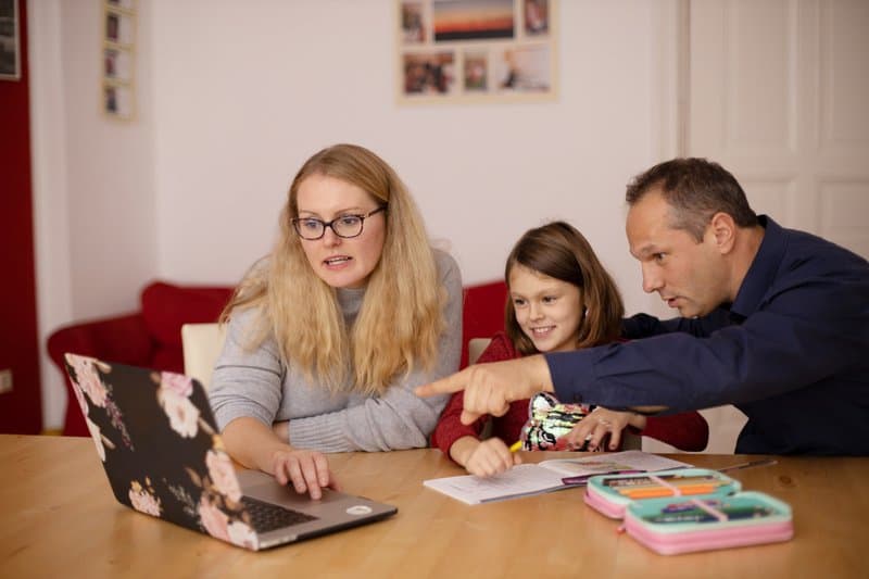 Een familie, een vader, moeder en hun jonge dochter, zit aan een houten tafel en kijkt samen naar een laptop met bloemenprint; de vader wijst naar het scherm terwijl de moeder typt en de dochter meevolgt, wat gezamenlijk leren en de rol van AI in gezinsverband illustreert.